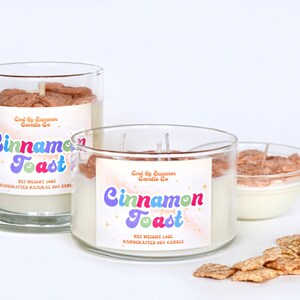May include: Two glass jars with white soy candles. The candles are topped with cinnamon toast cereal. The labels on the jars read "End of Summer Candle Co. Cinnamon Toast" and include the net weight of the candle. A small bowl of cinnamon toast cereal is in the background.