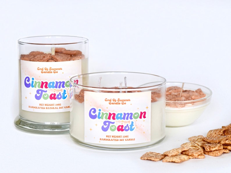 May include: Two glass jars with white soy candles. The candles are topped with cinnamon toast cereal. The labels on the jars read "End of Summer Candle Co. Cinnamon Toast" and include the net weight of the candle. A small bowl of cinnamon toast cereal is in the background.