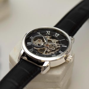 May include: A silver-toned skeleton watch with a black face and a black leather band. The watch face features Roman numerals and visible internal gears. The watch is displayed on a white surface.