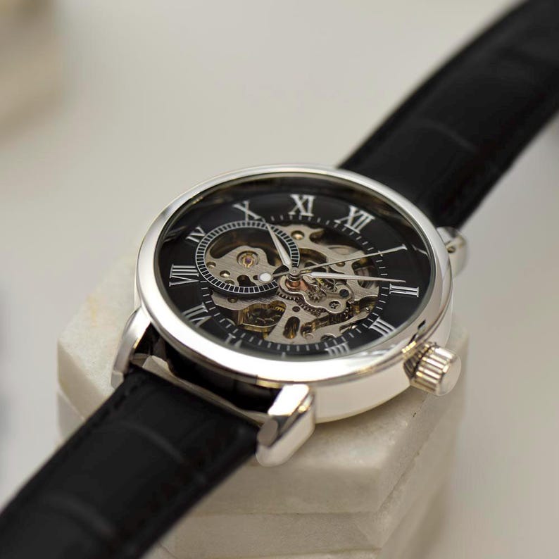 May include: A silver-toned skeleton watch with a black face and a black leather band. The watch face features Roman numerals and visible internal gears. The watch is displayed on a white surface.