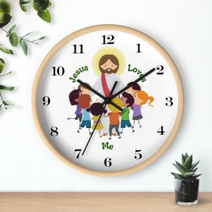 May include: A white round clock with a wooden frame features a colorful illustration of Jesus surrounded by children. The clock face has black Roman numeral hour markers and black hands. The text "Jesus Loves Me" is written in green letters around the illustration.