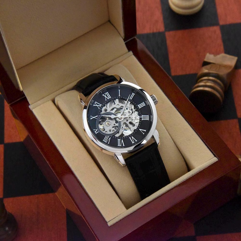 May include: A silver-toned skeleton watch with a black leather band is displayed in a wooden presentation box. The watch face features Roman numerals and visible internal gears. The box has a light tan interior and a polished, dark wood exterior.