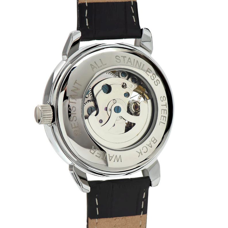 May include: Close-up of a silver-toned watch with a black leather band. The watch back is transparent, revealing the inner workings. The words "ALL STAINLESS STEEL", "WATER RESISTANT", and "BACK" are visible.