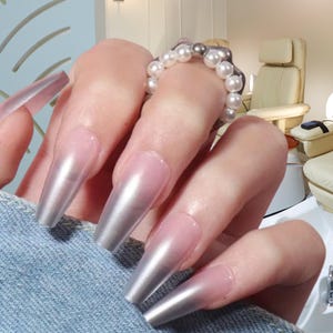 Op de afbeelding: Close-up van lange, zilverkleurige metallic acrylnagels. De nagels hebben een gradiënt, van zachtroze aan de basis tot glanzend zilver aan de uiteinden. Een parelring siert een vinger. De nagels zijn een schoonheids- en mode-uitspraak.