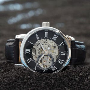 May include: A silver-toned mechanical watch with a black face and Roman numeral hour markers. The watch features a visible inner mechanism and a black leather band. The watch is displayed on a dark, textured surface.