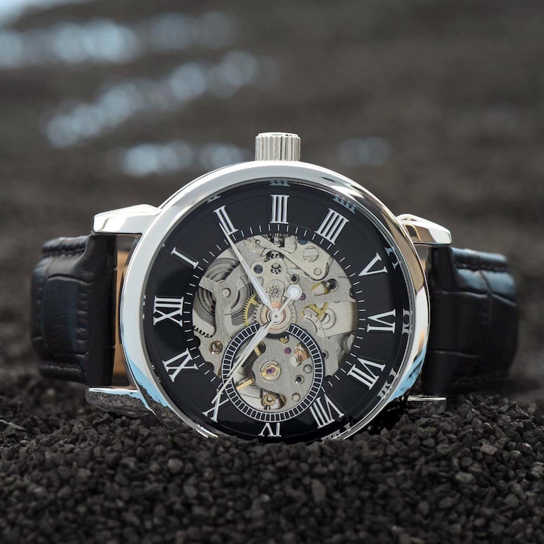 May include: A silver-toned mechanical watch with a black face and Roman numeral hour markers. The watch features a visible inner mechanism and a black leather band. The watch is displayed on a dark, textured surface.