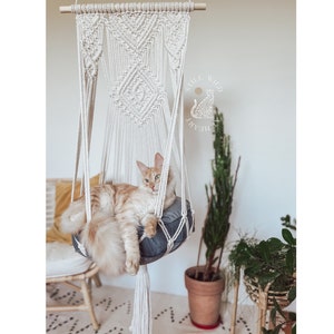 May include: A white macrame cat hammock with a grey cushion, hanging from a wooden dowel. A ginger cat is relaxing in the hammock. The text "Full Wild Pawsitive" is visible in the background.