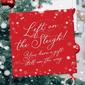 May include: A red square Christmas card with white script reading "Left on the Sleigh! You have a gift still on the way." The card is hanging on a Christmas tree with red ornaments and a small gold tree ornament.