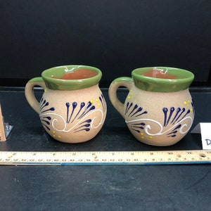 Op de afbeelding: Twee kleine, bruine keramische bekers met groene randen en blauwe en gele beschilderde ontwerpen. De bekers hebben een afgerond lichaam en een handvat.