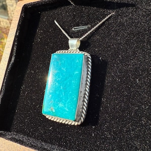 May include: A rectangular turquoise pendant with a silver-colored rope-like border and a silver chain. The bright blue-green stone is set in a silver frame, creating a striking contrast. The pendant is displayed on a black velvet surface.