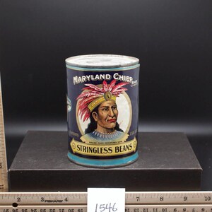 Op de afbeelding: Vintage blikje Maryland Chief Stringless Beans. Het blikje heeft een blauwe achtergrond met een rood-wit-blauwe afbeelding van een indiaanse opperhoofd met een verenhoed.