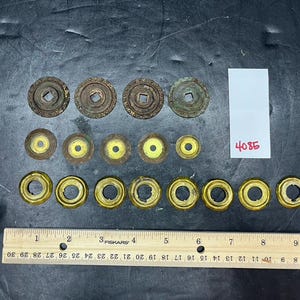May include: A collection of antique metal hardware components, featuring circular washers and decorative elements. A ruler with centimetre measurements is visible, along with the number "4085" on a white label.