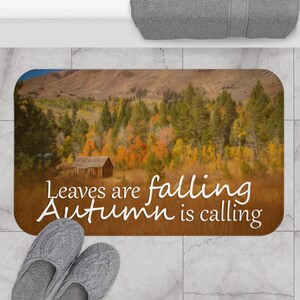 Op de afbeelding: Rechthoekige badmat met een herfstlandschap met een kleine houten hut, kleurrijke bomen en bergen. De mat heeft de tekst "Leaves are falling, Autumn is calling" in witte letters. Grijze pantoffels staan op de voorgrond.