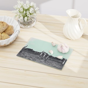 May include: A glass cutting board with a black and white image of birds on a lake. The board is on a light wood surface with garlic cloves scattered around it.