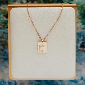 May include: A gold-toned necklace featuring a delicate chain and a small, rectangular pendant. The pendant is engraved with the text "CHILD OF GOD" in a clean, sans-serif font. The piece is displayed on a white, rectangular stand with gold edges.