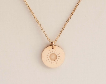 May include: A gold-toned necklace featuring a delicate chain and a circular pendant. The pendant displays a central circular cutout surrounded by engraved radial lines, creating a sunburst pattern on the smooth, polished metal surface.