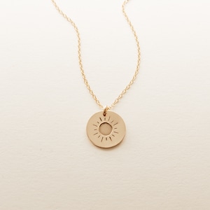 May include: Gold-colored necklace featuring a circular pendant with a sunburst design. The pendant is attached to a delicate chain. The necklace is displayed against a neutral background, highlighting the jewelry's simple elegance.