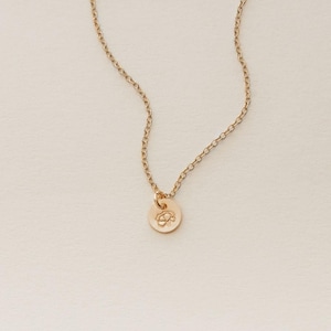 May include: A delicate gold-colored necklace with a small, round pendant. The pendant features a subtle floral design. The chain is a classic link style, and the necklace is displayed against a neutral background.