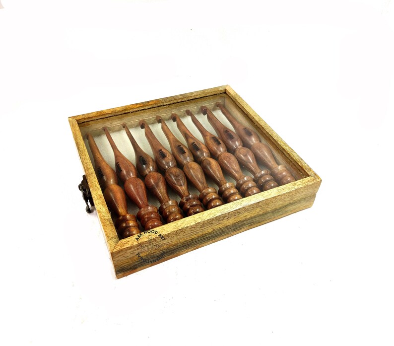 May include: A set of ten wooden crochet hooks in a wooden box with a clear lid. The hooks are arranged in a row and are all the same size. The box is made of dark wood with a light wood trim. The box has a small metal latch on the side.