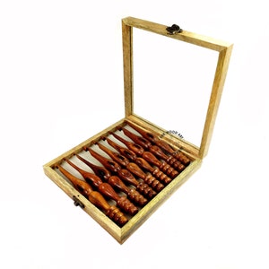 May include: A set of eight wooden crochet hooks in a wooden box with a glass lid. The hooks are different sizes and are arranged in the box in order of size. The box is made of light wood and has a brass latch.