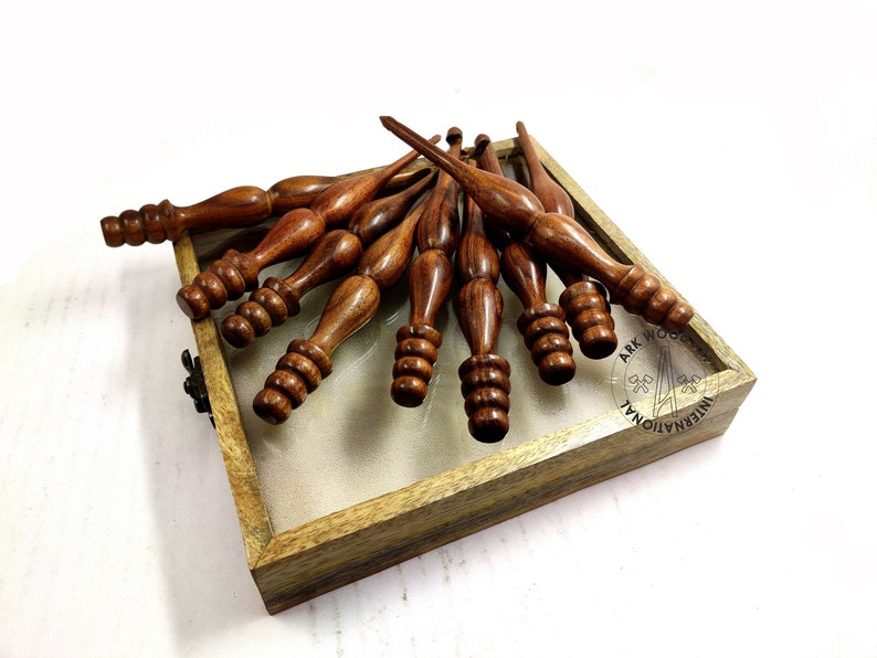 May include: A set of ten wooden knitting needles in a wooden box. The needles are brown and have a pointed tip. The box is made of light brown wood and has a clear glass top.