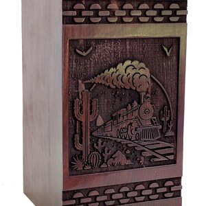 Peut inclure: Un coffret en bois avec un motif sculpté représentant un train, des cactus et un paysage désertique. La boîte est rectangulaire avec un côté clair et une face marron foncé avec des bordures décoratives. Le train émet de la fumée.