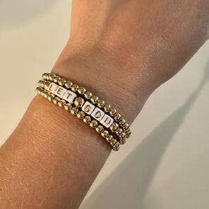 May include: Gold beaded bracelets stacked on a wrist. One bracelet features white letter beads spelling "LET GOD". The bracelets are made of small, round gold beads.