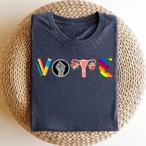 May include: Dark grey t-shirt with the word "VOTE" spelled out in colorful graphics. The letters are made up of a pencil, a fist, a uterus, and a rainbow.