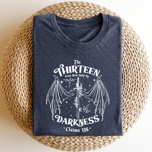 May include: A dark grey t-shirt with a white graphic design featuring a winged creature and a sword. The text "The Thirteen From Now Until The Darkness Claims Us." is printed on the shirt.