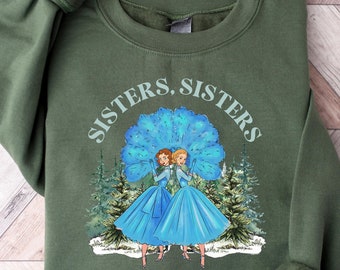 Puede incluir: Una sudadera verde oliva con el texto "Sisters, Sisters" sobre una ilustración de dos mujeres con vestidos azules. La ilustración está ambientada sobre un abanico azul y árboles de hoja perenne. El texto "There Were Never Such Devoted Sisters" está debajo.