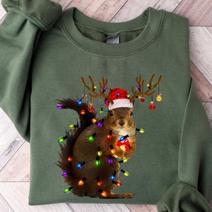 May include: Green sweatshirt featuring a festive squirrel design. The squirrel is adorned with a Santa hat, antlers, and Christmas lights. It holds a wrapped gift, creating a whimsical holiday theme. The sweatshirt is a solid green color.