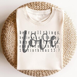 May include: A beige t-shirt with the text "bears all things, believes all things, hopes all things, endures all things. 1 Corinthians 13:7" printed on it. The word "love" is written in a large, cursive font in the center of the shirt.