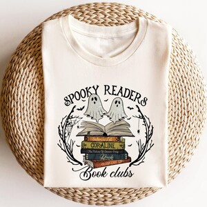 May include: Cream-colored t-shirt featuring a graphic design with the text "Spooky Readers Book Clubs." The design includes two ghost figures above a stack of books, with bats and spiderwebs. The books have titles like "Coraline" and "Frankenstein."
