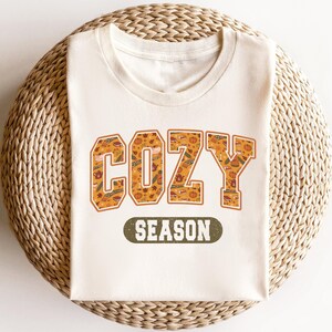 May include: A white t-shirt with a graphic print of the word "COZY" in orange and brown with a fall-themed pattern. The text "SEASON" is printed in green below the word "COZY".