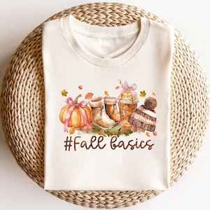 May include: A white t-shirt with a watercolor illustration of fall-themed items, including boots, a pumpkin, and a coffee drink. The text "#Fall Basics" is printed below the illustration.