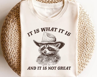 May include: A white t-shirt with a brown raccoon wearing a cowboy hat. The text on the shirt reads "It is what it is and it is not great."