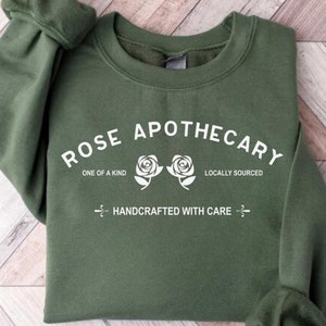 Puede incluir: Una sudadera verde con el texto "ROSE APOTHECARY" en blanco. El texto est&aacute; rodeado por dos rosas blancas y las palabras "ONE OF A KIND" y "LOCALLY SOURCED". Debajo del texto est&aacute; la frase "HANDCRAFTED WITH CARE".