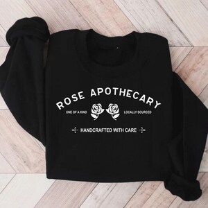 Puede incluir: Sudadera negra de manga larga con el texto "ROSE APOTHECARY" en blanco, con dos rosas blancas y el texto "ONE OF A KIND", "LOCALLY SOURCED" y "HANDCRAFTED WITH CARE" en blanco.