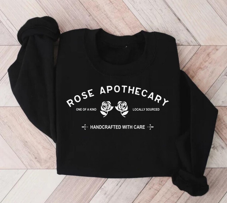 Puede incluir: Sudadera negra de manga larga con el texto "ROSE APOTHECARY" en blanco, con dos rosas blancas y el texto "ONE OF A KIND", "LOCALLY SOURCED" y "HANDCRAFTED WITH CARE" en blanco.