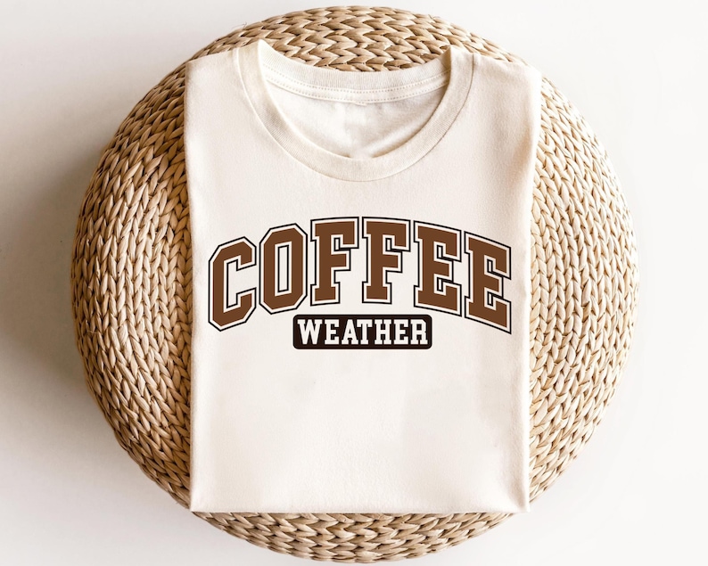 K&ouml;nnte beinhalten: Ein wei&szlig;es T-Shirt mit braunem Text, der "COFFEE WEATHER" lautet. Der Text ist in einem College-Schriftart-Stil.