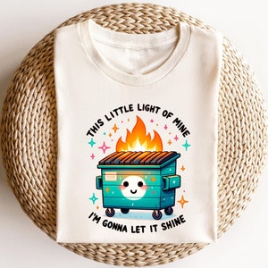 May include: A white t-shirt featuring a graphic of a green dumpster on fire with a smiling face. The text "This little light of mine" is above the dumpster and "I'm gonna let it shine" is below.