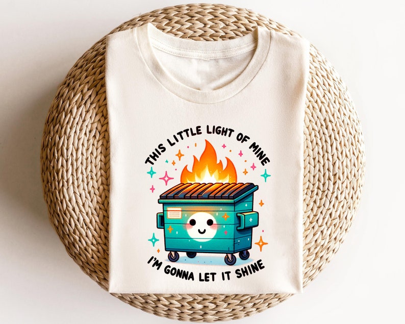 May include: A white t-shirt featuring a graphic of a green dumpster on fire with a smiling face. The text "This little light of mine" is above the dumpster and "I'm gonna let it shine" is below.