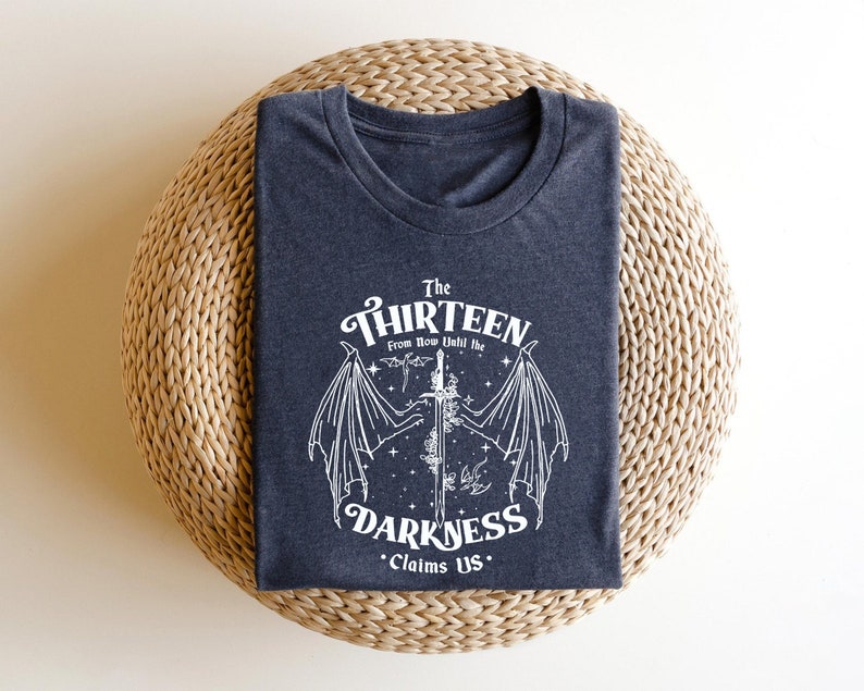 May include: A dark grey t-shirt with a white graphic design featuring a winged creature and a sword. The text "The Thirteen From Now Until The Darkness Claims Us." is printed on the shirt.