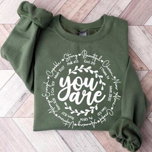 May include: A green sweatshirt with a white circle design that says "you are" surrounded by words and bible verses. The words include "strong", "beautiful", "chosen", "never alone", "capable", "enough", "amazing", "victorious", "always", and "greatly loved".