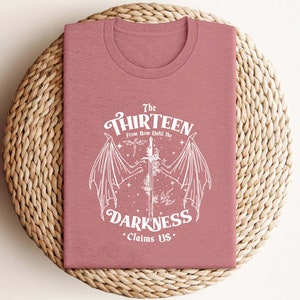 May include: A pink t-shirt with a white graphic design featuring a sword with wings and the text "The Thirteen From Now Until The Darkness Claims Us."