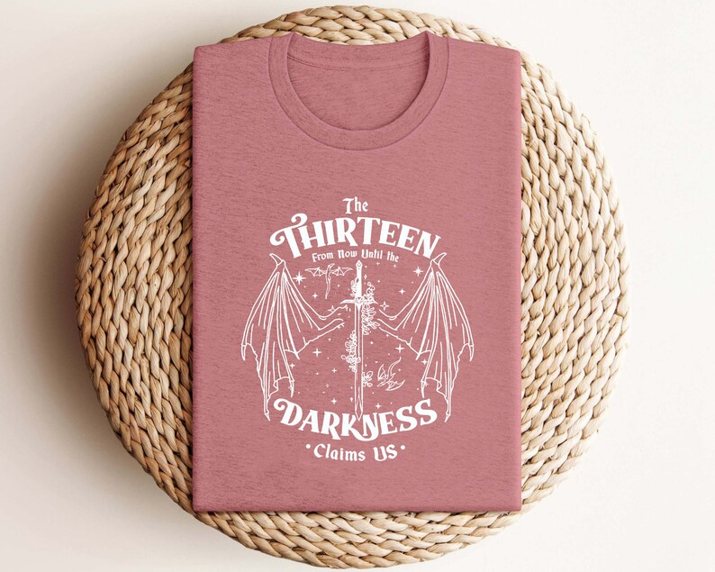 May include: A pink t-shirt with a white graphic design featuring a sword with wings and the text "The Thirteen From Now Until The Darkness Claims Us."