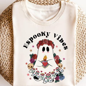 May include: Cream-colored t-shirt featuring a cartoon ghost with a floral crown, holding a heart with a flame. The ghost is surrounded by flowers, skulls, and the words "Espooky Vibes".