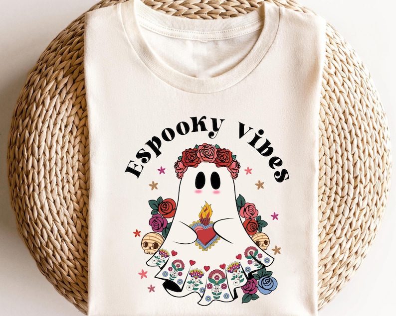 May include: Cream-colored t-shirt featuring a cartoon ghost with a floral crown, holding a heart with a flame. The ghost is surrounded by flowers, skulls, and the words "Espooky Vibes".