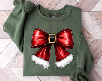 Puede incluir: Una sudadera verde oliva con un gran diseño de lazo rojo vibrante. El lazo tiene una hebilla dorada y un ribete de piel sintética blanca. La sudadera está hecha de un material suave, perfecta para la temporada navideña.