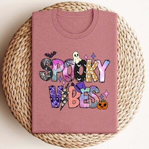 May include: A pink t-shirt with a colorful graphic that says "Spooky Vibes". The graphic includes a ghost, a witch's hat, a spiderweb, and a pumpkin.